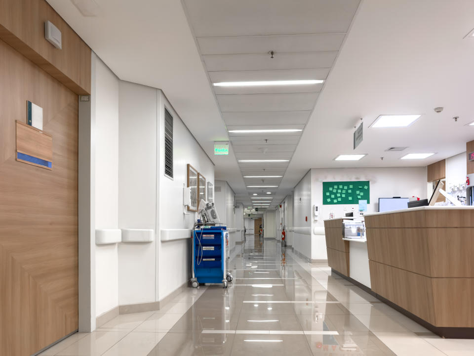 A hospital hallway with medical equipment, including a blue cart, is visible near a nurse's station. The corridor leads to multiple rooms and areas