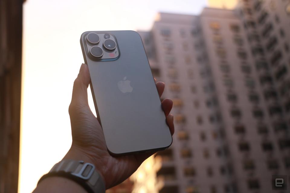The iPhone 15 Pro Max held up in mid-air with a New York building in the background while the sun sets.