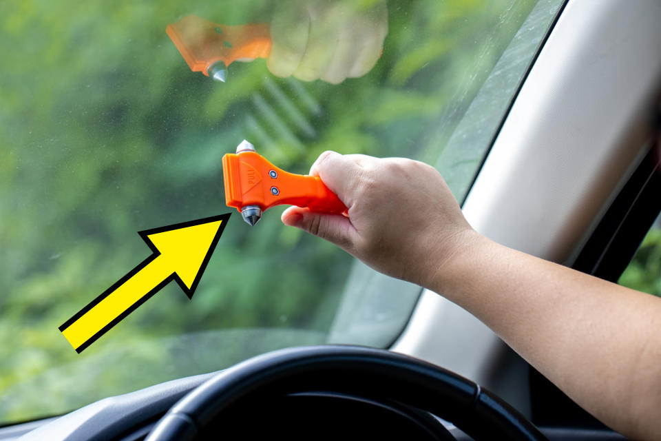 A hand is holding an orange emergency glass breaker tool against a car window, ready to break it. The car interior and steering wheel are partially visible