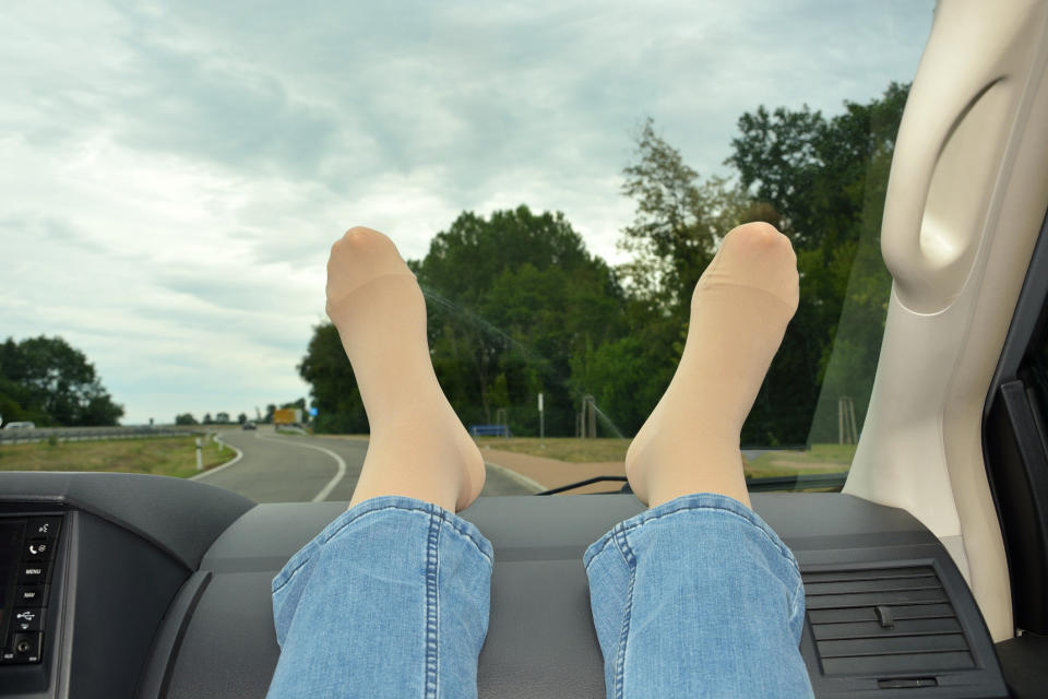 Person with legs extended onto a car dashboard, driving on a curvy road with trees and a cloudy sky visible through the windshield