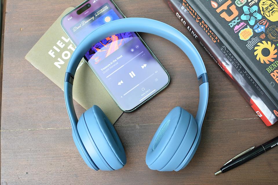 <p>Beats Solo 4 headphones laying on a wooden table with a notebook and an iPhone underneath.</p>
