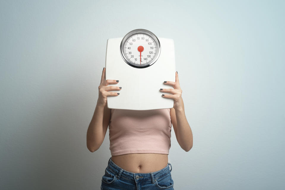 Young woman hides behind  bathroom scales. Studio shot. By emphasizing behaviors over the scale, people can avoid the disappointment linked to the uncontrollable nature of weight numbers. (Getty Images)