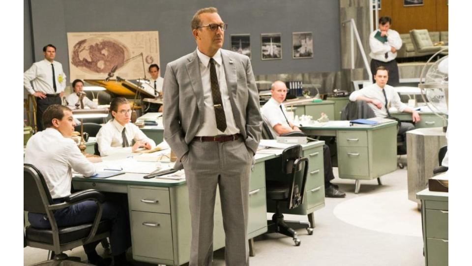 Kevin Costner in Hidden Figures; he stands in a suit while men sit at desks behind him.