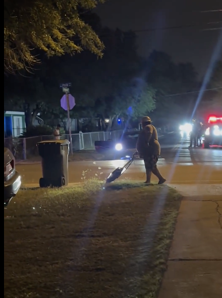 A person is sweeping leaves off the sidewalk at night in a suburban area. Cars with headlights on and one with flashing lights are visible in the background