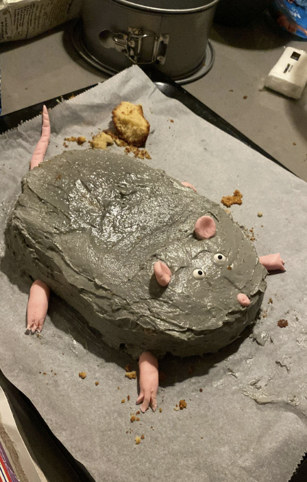 A cake shaped like a rat with grey frosting and fondant details on the face and legs, placed on parchment paper