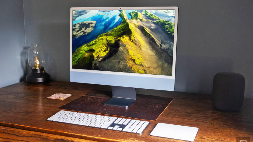 The 2023 Apple iMac with a 24-inch screen and the M3 chip in ice blue sits on a worn wooden corner desk next to a small lamp and an Apple HomePod.