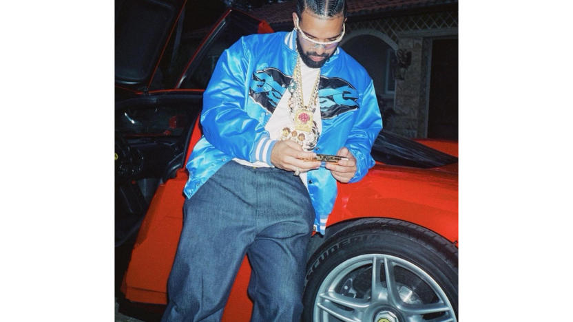 The rapper Drake leaning against a red car, looking at his phone.