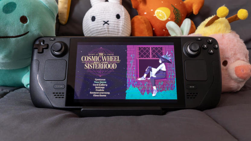 A Steam Deck sitting on a blanket with cute plush characters behind it. On its screen is The Cosmic Wheel Sisterhood’s title screen.