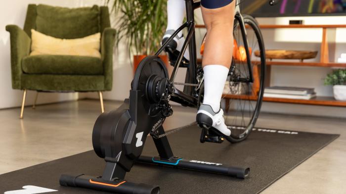 A Zwift Hub One bike trainer seen in use from behind. A person's bike is connected and indoors facing a TV with the Zwift app onscreen.