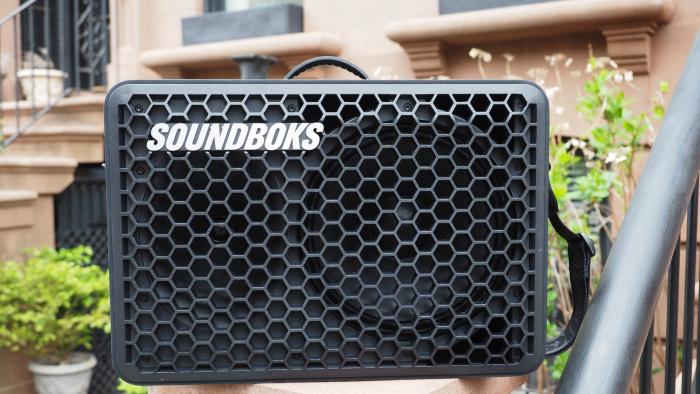 The Soundboks Go portable Bluetooth speaker sitting in front of a Brooklyn brownstone, with plants, stairs and railings in the background.