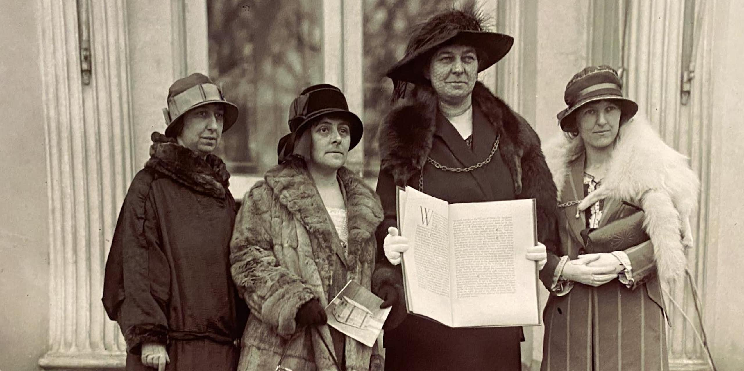 Four smartly dressed women with hats stand with a large book on the steps of the White House