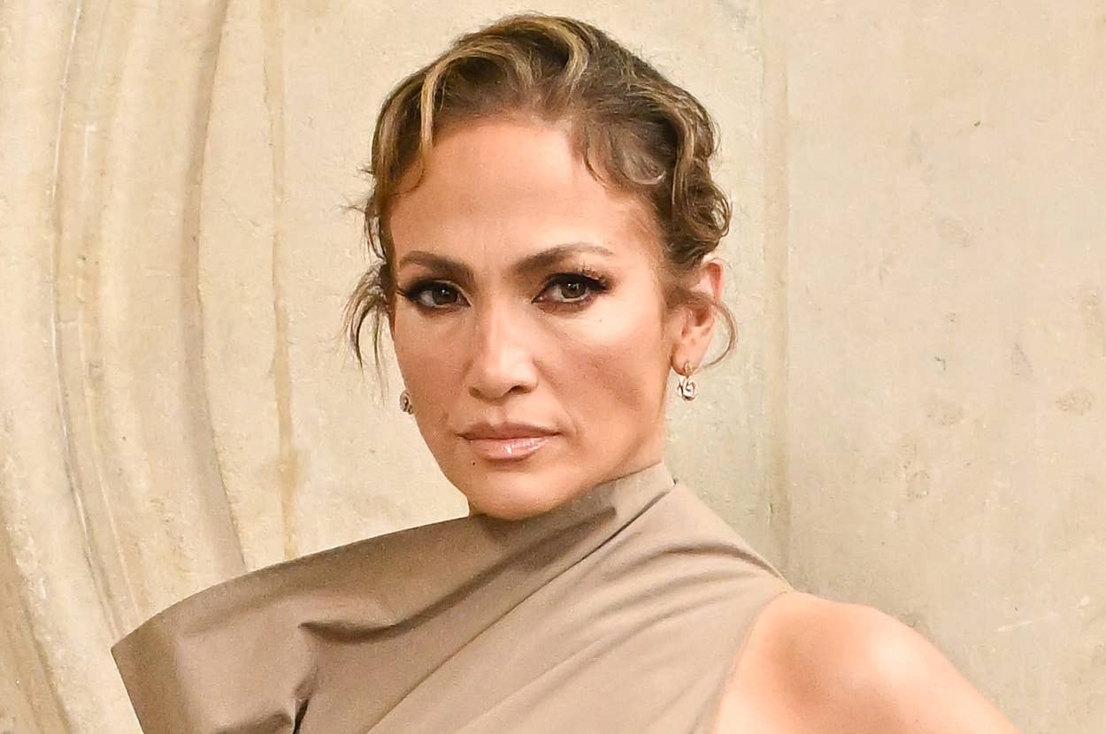 Jennifer Lopez poses against a stone wall, wearing a sleeveless, asymmetrical high-neck dress and black elbow-length gloves. Her hair is styled in an updo