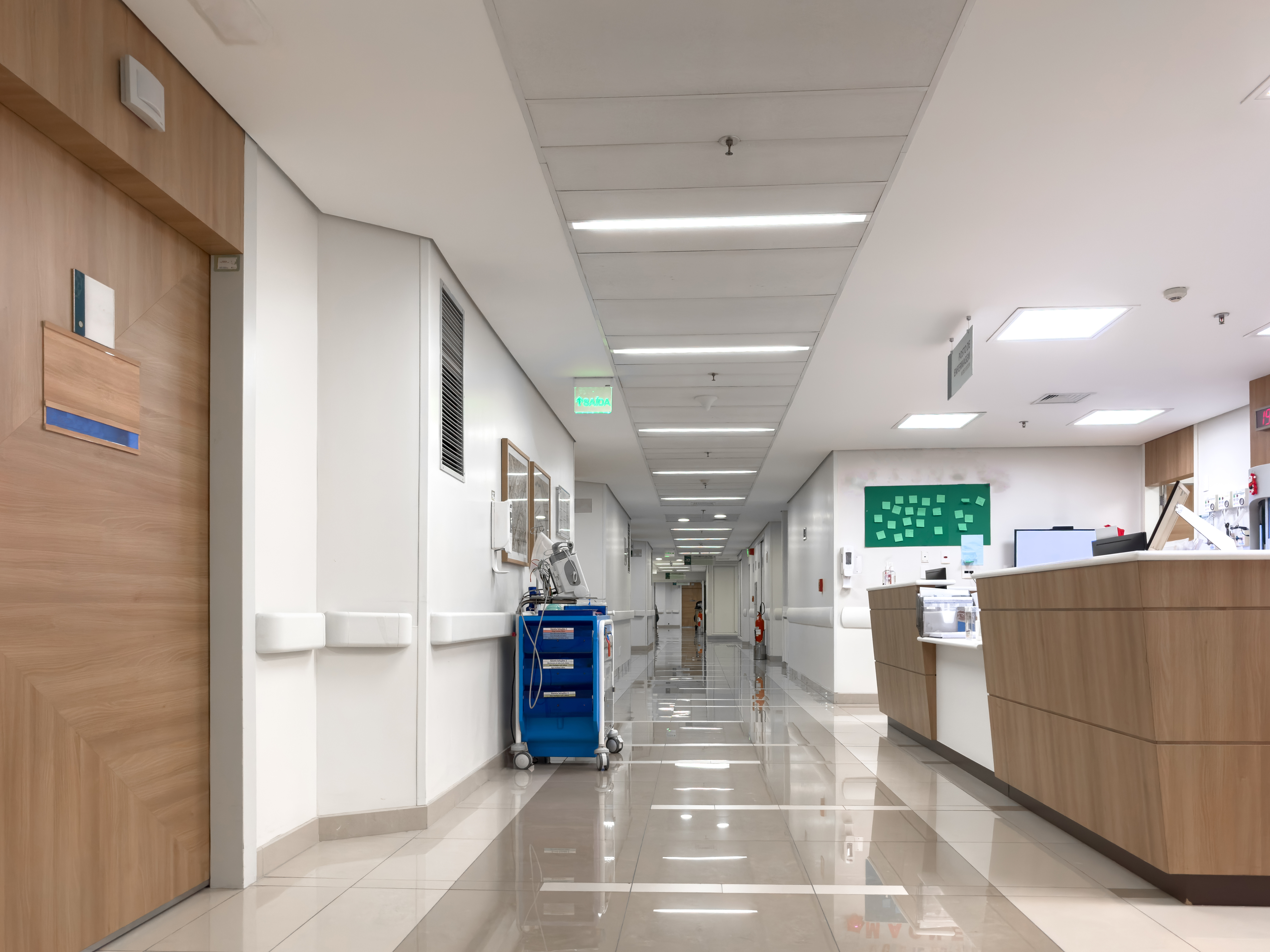 A hospital hallway with medical equipment, including a blue cart, is visible near a nurse&#x27;s station. The corridor leads to multiple rooms and areas